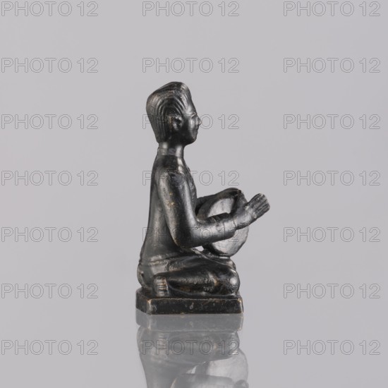 Musician figurine, art of Southeast Asia, Southeast Asia culture, musicians, 1st half of the 20th century contemporary product, Cambodia place of origin, sculpture, figurine, height 6.3 cm, A small dark statue of a seated figure viewed in profile, shown kneeling on a square base with hands raised and holding a rounded object, rendered with smooth surfaces and subtle carved hair texture, placed on a reflective surface; visible colors include black with hints of dark brown and a neutral gray background.