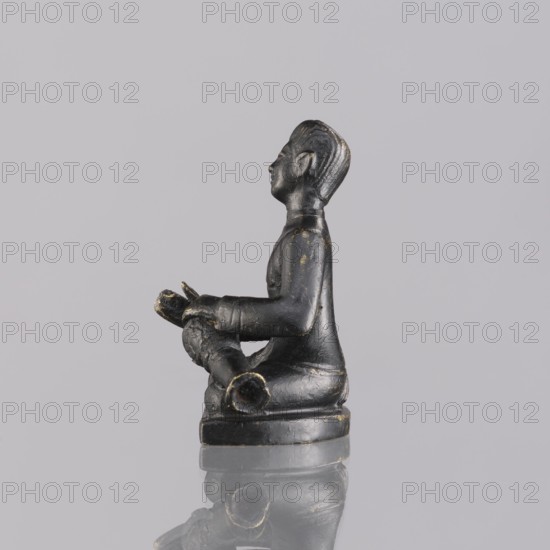 Musician figurine, Southeast Asian art, musicians, Southeast Asia culture, 1st half of the 20th century contemporary product, Cambodia, sculpture, figurine, height 6.3 cm, A small sculpted figure in a seated pose made of a dark material with worn golden highlights, shown in profile facing left, with a defined head, facial features, and molded hair, wearing a long-sleeved garment and seated on a rounded base; the figure holds a cylindrical object across its lap with visible texture and open ends, and the object and the figure show areas of wear revealing lighter tones; the scene rests on a reflective surface, and the background is a plain pale gray.