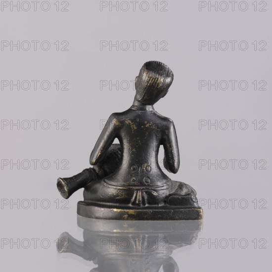 Musician figurine, Southeast Asian art, musicians, Southeast Asia culture, 1st half of the 20th century contemporary product, Cambodia, sculpture, figurine, height 6.3 cm, A small dark metallic figurine with worn gold highlights shows the back of a seated person on a rectangular base, with short textured hair and arms resting at the sides; a cylindrical object lies beside the figure and the base and figure are reflected on a smooth reflective surface against a pale gray background.