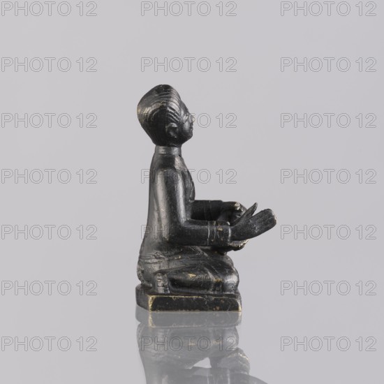 Musician figurine, Southeast Asian art, musicians, Southeast Asia culture, 1st half of the 20th century contemporary product, Cambodia place of origin, sculpture, figurine, height 6.3 cm, A small dark figurine of a seated person shown in profile on a reflective surface, with a muted light gray background; the figure appears black with worn areas revealing a slightly lighter, gold-tinged color, showing textured hair, defined facial features, a collar, long sleeves, and hands held forward, seated on a square base that also shows signs of wear and similar black and gold-tinged tones.