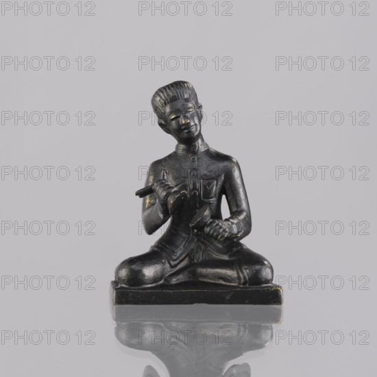 Musician figurine, Southeast Asian art, Southeast Asia culture, musicians, 1st half of the 20th century contemporary product, Cambodia place of origin, sculpture, figurine, height 6.5 cm, A small dark figurine with a shiny black surface sits cross-legged on a rectangular base, depicting a person holding a cylindrical object in one hand and a rounded tool in the other, with textured hair, a high-collared garment, and visible folds in the clothing; the scene shows reflections on the smooth surface beneath the base and the background is a pale gray.
