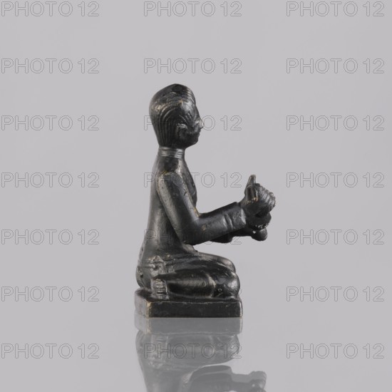 Musician figurine, creator unknown, Southeast Asian art, Southeast Asia culture, musicians, provenance unknown, 1st half of the 20th century contemporary product, Cambodia place of origin, sculpture, figurine, height 6.5 cm, A small dark statue of a kneeling human figure viewed in profile, sitting on a square base with a reflective surface beneath; the figure appears black with a few faint highlights of lighter wear and has textured hair, a defined face in profile, arms extended forward holding a small rounded object, and clothing indicated by smooth contours and slight folds; the background is a uniform light gray.