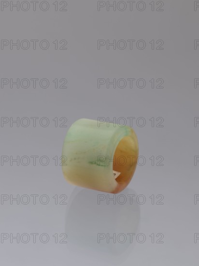 Ring, Chinese, Qing dynasty 1644–1911, green-brown jade ornament/jewelry, 18th–19th century, China; height 1.5 cm, diameter 2.0 cm., A smooth cylindrical ring with a rounded outer surface sits on a reflective surface, displaying a gradient of pale green to yellowish-orange hues with subtle marbling and a few faint darker streaks, against a plain soft gray background.