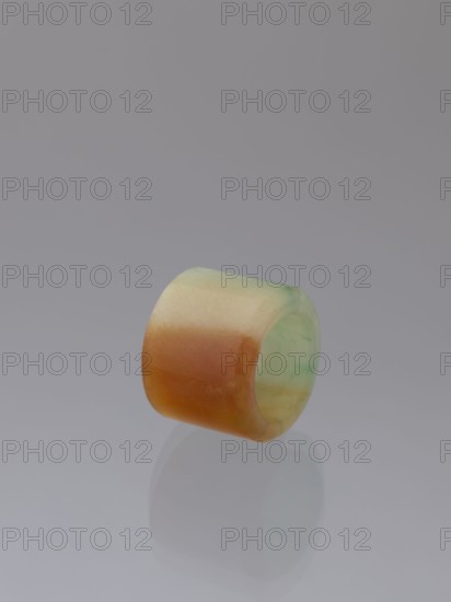 Ring, Chinese, Qing dynasty 1644–1911, green-brown jade ornament, 18th–19th century, China; jewelry; height 1.5 cm, diameter 2.0 cm., A cylindrical, hollow object with smooth, rounded edges rests on a reflective surface; its outer surface shows a gradient of colors including green, yellow, and orange, while the inner surface appears pale green with subtle marbling. The background is a uniform gray that transitions slightly in tone, and the object's reflection is visible beneath it on the glossy surface.