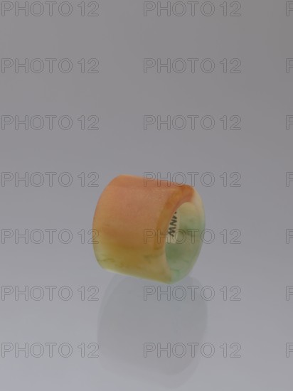 Ring, Chinese, Qing 1644–1911, green-brown jade, 18th–19th century, China; jewelry; height 1.5 cm, diameter 2.0 cm., A smooth cylindrical ring with a wide band sits on a reflective surface, showing a gradient of colors including orange, peach, yellow, pale green, and a hint of mint; the background is a uniform cool gray and the ring casts a soft reflected silhouette on the surface.