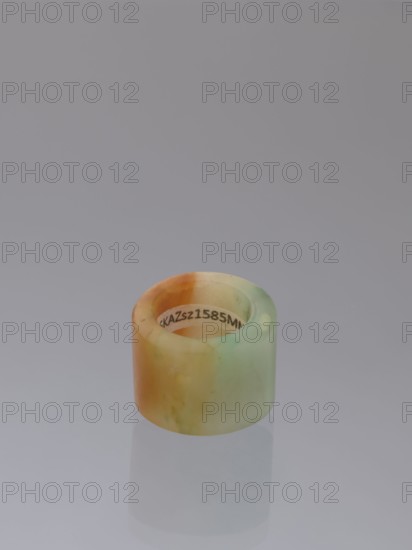Ring, Chinese, Qing 1644–1911, green-brown jade ornament/jewelry, 18th–19th century, China; height 1.5 cm, diameter 2.0 cm., A small cylindrical object with a smooth, slightly translucent surface sits on a reflective surface, showing a pale gradient of orange, yellow, green, and cream tones; a faint reflection of the cylinder appears below it, and the background is a uniform soft gray.