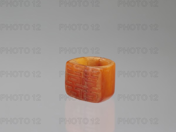 Signet ring, Chinese, Qing dynasty 1644–1911, 18th–19th century. Opaque amber with convex relief, ornament depicting the symbol for happiness. Jewelry; height 3.3 cm, width 3.1 cm, depth 2.6 cm., A small cylindrical object with a hollow center sits against a plain light gray background, showing shades of orange and amber with a slightly translucent quality; one face displays an array of carved rectangular grooves and shallow incised patterns while the opposite curved surface appears smooth and rounded, and a soft reflection of the object is visible on the glossy surface beneath it.
