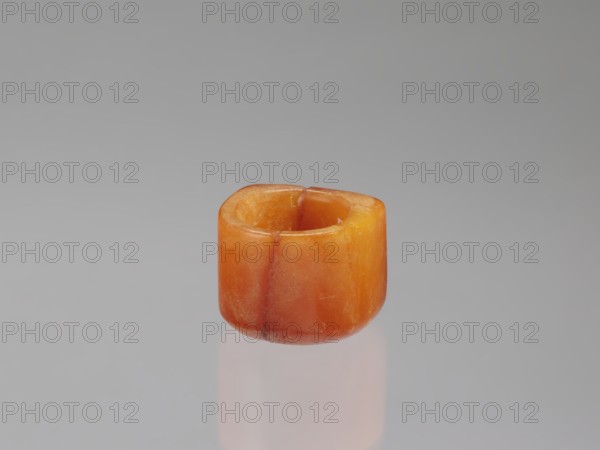 Signet ring, Chinese, Qing dynasty 1644–1911, opaque amber with convex relief, 18th–19th century, China; jewelry; ornament symbolizing happiness; height 3.3 cm, width 3.1 cm, depth 2.6 cm., A small cylindrical object with a rounded top and hollow center sits against a smooth neutral background, showing a glossy surface with visible seams and slight surface texture; the object displays a range of warm orange and amber tones with areas of deeper reddish-orange and lighter yellowish-orange, and a faint reflection of the object appears below it on the surface.