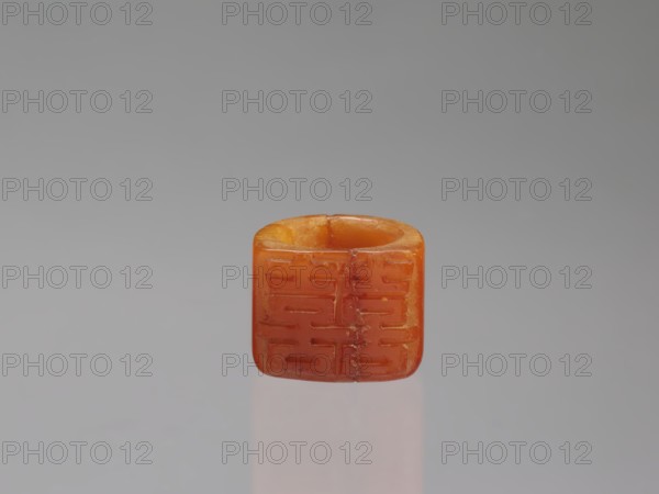 Signet ring, Chinese, Qing dynasty 1644–1911, opaque amber with convex relief, 18th–19th century, China; jewelry; ornament symbolizing happiness; height 3.3 cm, width 3.1 cm, depth 2.6 cm., An orange, square-shaped bead with rounded edges sits centered against a smooth gray background, showing a hollowed top and subtle surface grooves on its faces; the bead displays variations of orange and amber tones with small darker inclusions and a soft reflection beneath it on the surface.