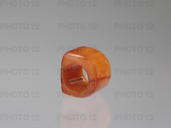 Signet ring, Chinese, Qing dynasty 1644–1911, 18th–19th century. Opaque amber with convex relief depicting a happiness symbol; jewelry. Dimensions: height 3.3 cm, width 3.1 cm, depth 2.6 cm., An orange, rounded rectangular ring with a smooth, slightly translucent surface rests on a reflective gray surface; the ring shows variations of orange with darker orange edges and lighter orange highlights, and there is a shadowed reflection beneath it.