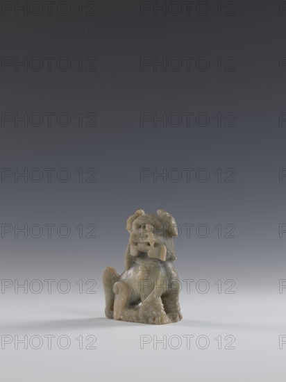 Seated lion, Chinese, lion fo iconography, Qing dynasty 1644–1911. Figurine carved in soapstone elephantine, engraved. 18th–19th century. Sculpture/figurine. Height 10.5 cm; width 7.5 cm; depth 4.0 cm., A small carved figurine made of a greenish-gray stone sits on a smooth light-gray surface against a dark gray gradient background; the figurine depicts a seated creature with a rounded body, curled tail, prominent snout, open mouth, stylized limbs, and textured surface, showing variations of green, gray, and subtle brown tones.