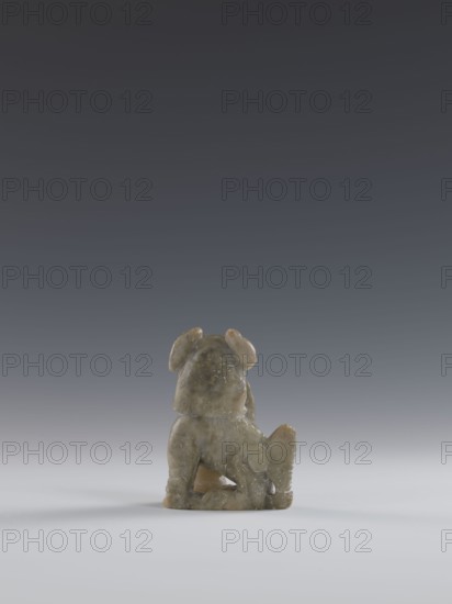 Seated lion, Chinese, lion fo iconography, Qing dynasty 1644–1911. Figurine carved in soapstone elephantine, engraved. 18th–19th century. Sculpture/figurine. Height 10.5 cm, width 7.5 cm, depth 4.0 cm., A small carved figure of an animal viewed from the back, made of mottled greenish-gray stone with pale brown and beige areas, sitting on a smooth light-gray surface against a gradient dark-gray to medium-gray background, showing rounded ears, a short tail, and textured carved details on its body.