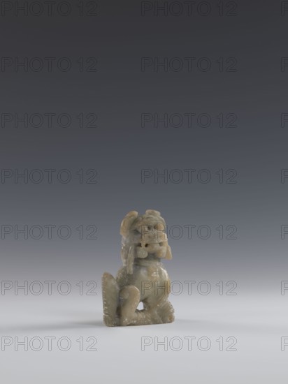 Seated lion, Chinese, lion fo iconography, Qing dynasty 1644–1911; soapstone elephantine carved and engraved figurine, 18th–19th century; sculpture/figurine; H 10.5 cm, W 7.5 cm, D 4.0 cm., A small carved figurine sits on a smooth surface against a gradient background. The figurine appears to be made of a muted greenish-beige stone with lighter and darker mottling. It has a rounded head with pronounced facial features and a prominent open mouth, flanked by curled elements that hang down beside the face. The body is compact with a curved back and folded limbs, and textured detailing suggests fur or scales along parts of the form. The surface shows subtle variations in sheen and some pitted or weathered areas. The scene includes colors of gray in the background, white in the foreground surface, and greenish-beige in the figurine.