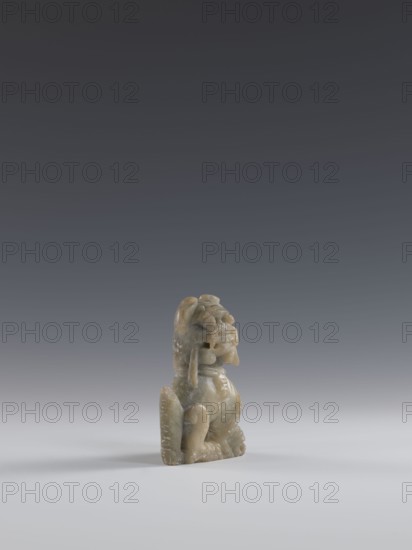 Seated lion, Chinese, Qing dynasty 1644–1911. Figurine carved in soapstone elephantine, engraved. 18th–19th century. China. Sculpture, figurine; height 10.5 cm, width 7.5 cm, depth 4.0 cm. Iconography: lion fo., A small carved figurine sits on a smooth surface against a gradient background. The figurine appears to be made of a mottled greenish-gray stone with lighter and darker patches. It has a rounded head with carved facial features and a pronounced open mouth, flanked by rounded ear-like shapes and a pair of hanging elements at the sides. The body is compact with curved limbs and a seated posture, showing smooth polished areas and some rougher textured spots. The surface of the base and parts of the figure show faint pale speckling. The foreground surface is a pale gray, transitioning into a darker gray backdrop that becomes almost charcoal toward the top of the image.