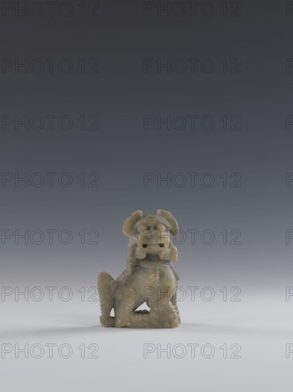 Seated lion, Chinese, lion fo iconography, Qing dynasty 1644–1911. Carved soapstone elephantine, engraved; figurine sculpture, 18th–19th century, China. Dimensions: height 10.5 cm, width 7.5 cm, depth 4.0 cm., A small carved figurine sits centered on a smooth surface against a gradient background; the figurine is made of a mottled greenish-gray material with lighter and darker patches and a slightly translucent quality, and it depicts a stylized animal with rounded ears, a pronounced snout, a curled tail, and textured surfaces that suggest fur or scales, with a worn, slightly rough finish and subtle highlights on raised areas while the surface beneath is pale gray and the background shifts from light gray near the bottom to dark gray at the top.