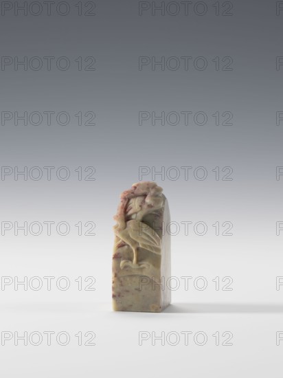 Seal stamp, Chinese, Qing 1644–1911, 19th–20th century. Blind seal stamp with square cross-section, height 6.9 cm, width 2.7 cm, depth 2.7 cm. Decoration: cranes in bas-relief and engraving., and provenance., A small carved stone object stands upright on a flat white surface against a gradient gray background; the stone has a warm beige base color with veins and patches of reddish and brown tones, and its carved face shows a raised bird motif perched on a curved element with stylized organic shapes above and around it, the carving exhibiting smooth and rounded contours and subtle shadowing.
