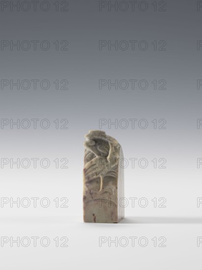 Seal stamp, Chinese, Qing 1644–1911, unknown provenance. Subject: cranes iconography. Seal stamp, blind, square cross-section. Decoration: cranes in bas-relief and engraving. 19th/20th century, China. Dimensions: height 6.9 cm; width 2.7 cm; depth 2.7 cm., A carved stone object stands upright on a flat surface, showing a sculpted relief of a bird with a curved neck and beak perched among stylized foliage near its top; the stone displays mottled tones of pale green, beige, and brown with some darker brown spots and streaks, and it casts a soft shadow to one side against a smooth light gray to white gradient background.