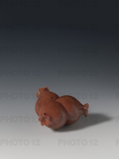 Three connected pomegranates fragment of a whole., Chinese; Qing dynasty 1644–1911. Object in the shape of three connected pomegranates, each with small holes on both sides; fragment of a whole. 18th–19th century, China. Sculpture pendant/varia. Height 4.8 cm; width 8.2 cm. Pomegranates fruits., A group of three reddish-brown, round objects with glossy surfaces and star-shaped protrusions at their tips resting together on a smooth, light gray surface, casting a soft shadow, set against a background that transitions from light gray near the bottom to dark gray toward the top.