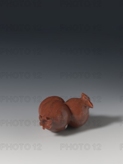 Three connected pomegranates fragment of a whole, Chinese, Qing 1644–1911, pomegranates fruits, pendants. Object in the shape of three connected pomegranates; each has small holes on both sides. Fragment of a whole. 18th–19th century, China. Sculpture, varia. Height 4.8 cm; width 8.2 cm., A group of three rounded, reddish-brown objects with matte surfaces sit together on a smooth, light gray surface, casting a soft shadow to the right; the background transitions from light gray near the bottom to dark gray and nearly black toward the top, and each rounded object has a small star-shaped protrusion at one end with subtle variations in reddish-brown tones and faint darker spots.