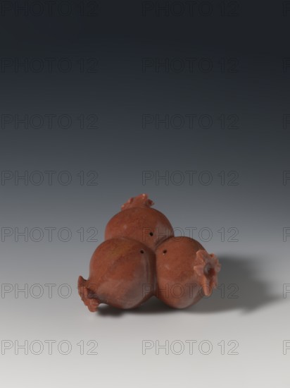 Three connected pomegranates fragment of a whole., Chinese. Qing dynasty 1644–1911. Object in the shape of three connected pomegranates; each has small holes on both sides. Fragment of a whole. Subject: pomegranates fruits; function: pendant. 18th–19th century. China. Sculpture. Varia. Height 4.8 cm; width 8.2 cm., A group of three rounded objects with textured surfaces and small protruding crowns, all in shades of reddish brown, sit together on a smooth gradient surface that shifts from light gray at the bottom to dark gray at the top; the objects cast soft gray shadows and show subtle variations of darker and lighter reddish brown across their curved forms.