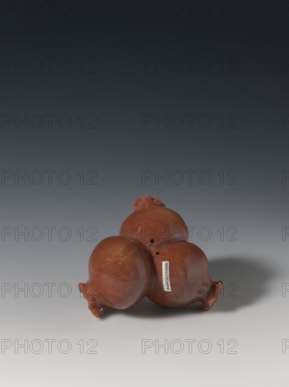 Three connected pomegranates fragment of a whole., Chinese; Qing dynasty 1644–1911. Object in the shape of three connected pomegranates, each with small holes on both sides; fragment of a whole. 18th–19th century. Sculpture; material/varia. Height 4.8 cm, width 8.2 cm. Pomegranates fruits; pendants., A group of three round, pomegranate-shaped objects clustered together, each with a rough textured surface and small crown-like protrusions; the objects are reddish-brown with areas of darker and lighter brown and a subtle glossy sheen. They sit on a smooth surface that transitions from light gray near the foreground to darker gray toward the background, and a soft shadow falls to the right of the cluster. The background above the surface fades from medium gray to a deep nearly black gradient.