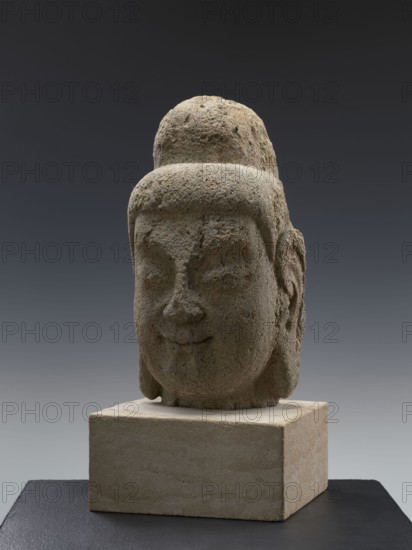 Head of Buddha, sculptor, Chinese; culture: Chinese; dynasties/styles: Qing 1644–1911, Song 960–1279; subject/iconography: Buddha ca. 560–480 BCE, ushnisha; Song-style sculpture; dated 19th/20th century; place of origin: China; medium: sculpture; height: 38.0 cm, A weathered stone sculpture of a head sits on a square stone base against a gradient dark gray to lighter gray background, showing a rounded topknot and stylized hair. The face is rounded with closed or softly downcast eyes, a broad nose, and a subtle smiling mouth; ears with carved inner details are visible at the sides. The stone surface is textured and pitted, with colors of gray, beige, and muted brown throughout, and the base shows sandy beige tones while the supporting surface below is dark gray to black.