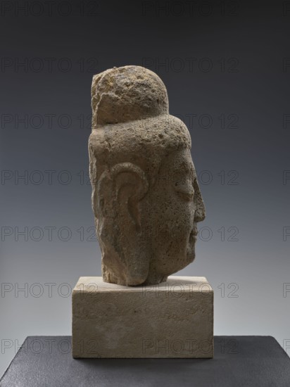 Head of Buddha; creator, provenance, Chinese; Qing 1644–1911; Song 960–1279 style; Chinese Buddhism; Buddha iconography Buddha ca. 560–480 BCE; ushnisha; Song-style sculpture; 19th/20th century; China; sculpture; height 38.0 cm, A weathered stone head sculpture shown in profile on a rectangular stone base against a smooth gradient background, with visible textures of rough, pitted stone and softer worn areas; the sculpture displays rounded hair or head covering, a defined ear, closed eye, and a slightly projecting nose and lips. The colors visible include various shades of beige, tan, brown, gray, and muted ochre on the sculpture and base, with a gradient background ranging from dark gray at the top to lighter gray toward the bottom and a dark gray surface beneath the base.