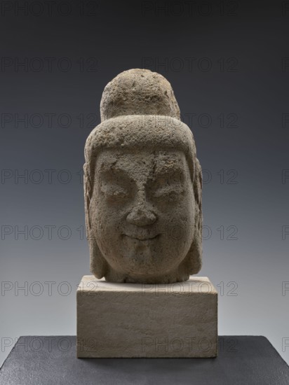 Head of Buddha, unknown sculptor; Chinese; Song-style sculpture; materials: sculpture; date: Song 960–1279 and Qing 1644–1911 influences / 19th–20th century; subject: Buddha iconography, ushnisha; dimensions: height 38.0 cm; cultural context: Chinese Buddhism; approximate original Buddha date referenced ca. 560–480 BCE., A weathered stone sculpture of a human head sits on a rectangular stone base, showing a rounded topknot and smooth, rounded facial features with closed eyes and a faint smiling mouth; the stone surface is rough and pitted with textured granularity. The colors visible include shades of gray, beige, and muted brown on the sculpture and base, with a gradient dark gray to lighter gray background and a dark slate-gray surface beneath the base.