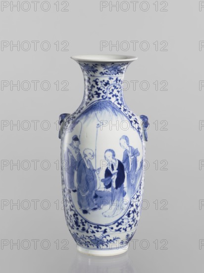 Vase, Chinese, Qing dynasty Kangxi period, 1662–1722. Blue-and-white porcelain ovoid vase with a flaring cup-shaped mouth. Shoulders with two applied mascarons with wheel motifs. Underglaze cobalt-painted decoration: figural scenes in two quatrefoil reserves; background plant ornament with small leaves and large floral rosettes, lotus tendril and lotus motifs, fans. Inscribed/signed. Vessel; height 25.5 cm, diameter 11.1 cm., A tall ceramic vase with a flared rim, shown against a plain light gray background, predominantly white with detailed blue painted decoration; the neck and lower body feature scrolling floral and foliate patterns in blue, and the main body has an oval panel painted in blue depicting a group of figures standing under a curved canopy; the vase has a pair of small sculpted handles on the shoulders glazed in blue, and the base area shows additional blue floral motifs.