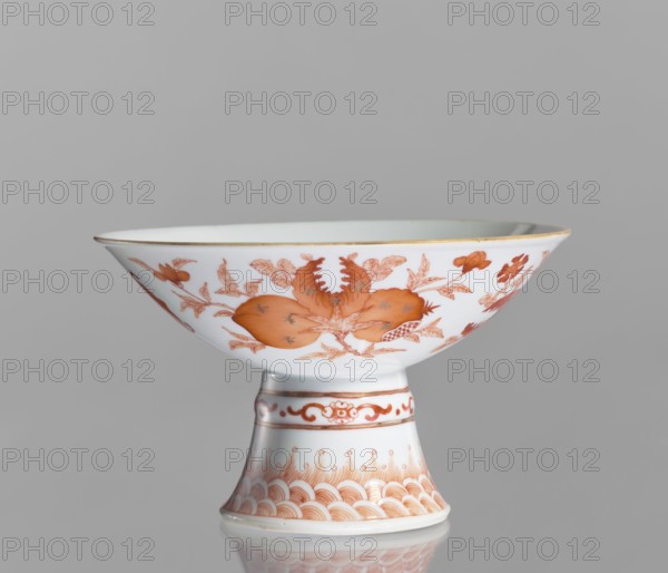 Platter, Chinese, ceramics; colored porcelain decorated with pomegranates, bats, peaches, and floral motifs symbolizing happiness. Porcelain platter on a tall, widening foot with white glaze. Decoration painted in red overglaze iron: center shows a peach, pomegranate, and Buddha's hand in a centripetal composition; exterior has three flowered branches bearing peach, pomegranate, and Buddha's hand, a sign of happiness surrounded by four bats; ornamental bands on the foot. 19th century, China. Vessel; height 11.7 cm, diameter 19.5 cm., A white porcelain stemmed bowl with a flared rim and a tapered foot, decorated with orange botanical and fruit motifs and delicate orange ornamental patterns on the stem and base; the rim has a thin metallic-looking band, and the piece sits on a reflective gray surface against a plain light gray background.