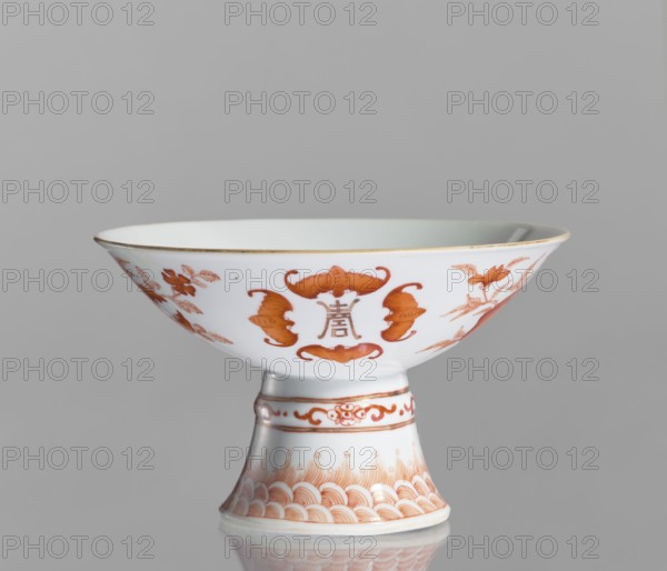 Platter, Chinese, ceramics. Porcelain platter on a tall, widening foot. Decoration painted with red overglaze iron: inside in the center centripetal composition a peach, pomegranate, and Buddha's hand; outside three flowered branches with peach fruit, pomegranate fruit, and a Buddha's hand, a sign of happiness surrounded by four bats; ornamental bands on the foot. White glaze. 19th century, China. Vessel; height 11.7 cm, diameter 19.5 cm., A white porcelain stem bowl with orange-red painted motifs sits against a gray background, showing a wide shallow bowl with a flared rim and a tapered pedestal foot; the bowl exterior features symmetrical ornamental shapes and stylized floral and bird-like designs in orange-red, with a thin gold rim at the top, while the pedestal displays bands of decorative orange-red patterns including wave-like motifs and scrollwork, and the piece casts a soft reflection on the surface below.