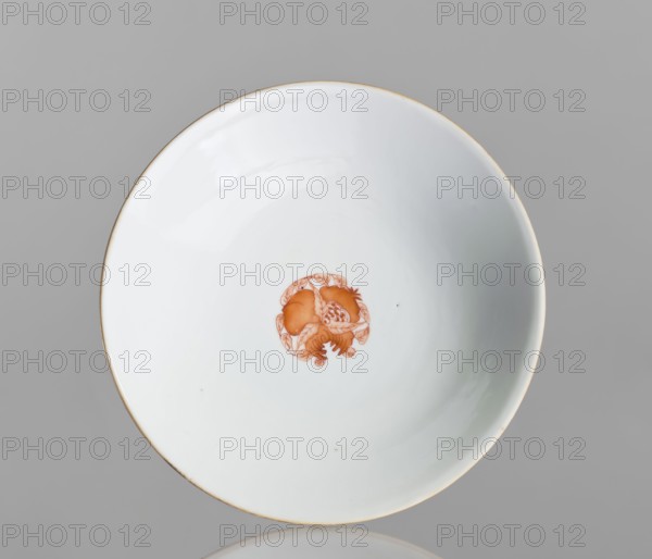 Platter, Chinese, 19th century. Porcelain platter on a tall, widening foot, white glaze, height 11.7 cm, diameter 19.5 cm. Decoration painted with red overglaze iron: inside center centripetal composition a peach, pomegranate, and Buddha's hand; outside three flowered branches with peach fruit, pomegranate fruit, and a Buddha's hand — a sign of happiness surrounded by four bats; ornamental bands on the foot. Motifs: pomegranates, bats, peaches, floral decorations; colored porcelain; symbol of happiness., A white shallow ceramic bowl with a thin gold rim is shown from above against a gray background, with a small orange decorative motif in the center and a few tiny dark specks on the interior surface; visible colors include white, gold, orange, gray, and small dark accents.