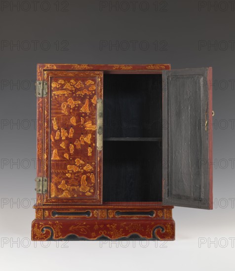 Cabinet, gift; figurative motifs; Chinese art; Chinese culture; furniture decorated with raised relief: figures in a landscape and plant motifs; courtly and genre scenes; floral motifs. Double doors; cabinet on a plinth cut out at the bottom in the outline of volutes; small shelf in the middle. Fittings and hinges made of brass sheet. Red, black and gold lacquer. 1st half of the 20th century, China. Piece of furniture. Height 50.0 cm; width 37.0 cm; depth 17.1 cm., A wooden cabinet with two doors, one closed and one open, decorated with painted scenes and patterns in red and gold on the exterior and a dark interior; the left door shows intricate gold-painted figures and landscape motifs on a red ground with metal hinges and a vertical metal latch plate, while the right door is opened to reveal a black-painted interior with a horizontal shelf and a textured inner panel; the cabinet sits on a carved base also painted red with gold floral and scroll motifs and areas of black accent, set a...