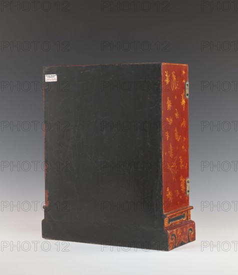 Cabinet, Decorated with raised relief: figures in a landscape on double doors; remaining surface decorated with plant and floral motifs and figurative courtly and genre scenes. Cabinet on a plinth cut out at the bottom in the outline of volutes; small shelf in the middle. Fittings and hinges made of brass sheet. Red, black and gold lacquer. 1st half of the 20th century, China. Piece of furniture. Height 50.0 cm, width 37.0 cm, depth 17.1 cm., A rectangular upright wooden box with a large flat front surface painted black showing visible scratches and worn edges, a decorative side painted red with gold floral and foliate motifs and small hinge hardware, and a red and gold ornamental base with scroll-like carved details; the background is a smooth gradient of dark gray to light gray and the surface beneath the box is a pale off-white, with visible colors including black, red, gold, gray, and off-white.