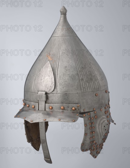 Helmet, 16th century, Turkey; Islamic, Turkish, Ottoman dynasty 1299–1922. Militaria headgear with movable cheekpieces and a nosepiece passing through the browband. Fluted bell. Browband and cheekpieces connected with copper rivets; neckpiece additionally with brass hinges. Weight 1.92 kg; height 39.0 cm; width 37.0 cm; depth 24.0 cm., A metal conical helmet with a pointed finial at the top, showing a weathered silver-gray surface with areas of darker gray patina and small patches of copper-colored wear; the helmet has vertical ridged decorations on the dome and a teardrop-shaped raised plate on the front, a brim that angles down slightly at the front, and a chin strap hanging below; a band around the lower dome is secured with a group of round copper-colored rivets, and additional copper-colored rivets decorate the ear guards and lower edges, which are segmented and show inner brownish leather lining.