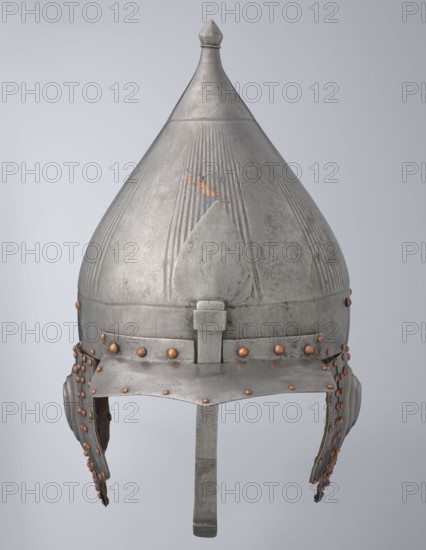 Helmet, Turkish, Ottoman dynasty 1299–1922, 16th century. Militaria headgear with movable cheekpieces and a nosepiece passing through the browband. Fluted bell. Browband and cheekpieces connected with copper rivets; neckpiece additionally with brass hinges. Weight 1.92 kg; height 39.0 cm; width 37.0 cm; depth 24.0 cm., A metal conical helmet with a pointed finial on top, showing a textured surface with vertical grooves and areas of wear, primarily silver-gray in color with patches of a warmer coppery-brown where the finish is worn and around a group of small round rivets; the helmet has a reinforced brow band and a central nose guard extending downward, along with cheek guards attached at the sides that hang slightly and are fastened by rows of coppery rivets, all set against a pale neutral background.