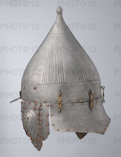 Helmet, Islamic art, Turkish, Ottoman dynasty 1299–1922, militaria headgear. Movable cheekpieces and a nosepiece passing through the browband. Fluted bell. Browband and cheekpieces connected with copper rivets; neckpiece additionally with brass hinges. 16th century, Turkey. Weight 1.92 kg; height 39.0 cm; width 37.0 cm; depth 24.0 cm., A metal helmet with a pointed conical top and a rounded dome featuring vertical ridged textures, mostly silver-gray in color with areas of darker gray patina, fitted with a lower brim and cheek guards. The surface shows numerous small rounded rivets in a coppery brown color arranged along edges and panels. There are a few small brass-colored metal fittings and hinges connecting parts of the helmet, including a few strap-like fasteners. One cheek guard has a curved shape with perforations and is attached by rows of coppery rivets. The overall object appears worn with slight surface scratches and variations in the silver-gray finish, set against a pale gra...