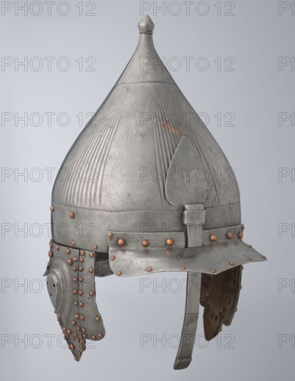 Helmet, Islamic art; Turkish, Ottoman dynasty 1299–1922; militaria headgear. Movable cheekpieces and a nosepiece passing through the browband. Fluted bell. Browband and cheekpieces connected with copper rivets; neckpiece additionally with brass hinges. 16th century, Turkey; weight 1.92 kg; height 39.0 cm; width 37.0 cm; depth 24.0 cm., A metal conical helmet with a pointed finial at the top, showing vertical ribbed texture on the upper sides and a smooth central panel, with a hinged nose guard and a downward curved brim; attached cheek guards hang on either side, each reinforced with a pattern of rounded rivets, and the metal surface shows areas of wear and small patches of brownish discoloration; visible colors include silvery gray metal, brownish spots, and coppery rivets against a neutral pale gray background.