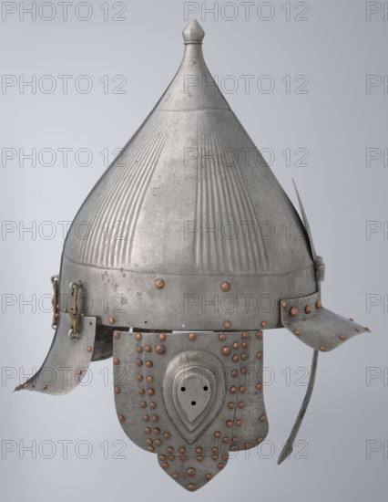 Helmet, Islamic art, Turkish, Ottoman dynasty 1299–1922, militaria headgear. Movable cheekpieces and a nosepiece passing through the browband. Fluted bell. Browband and cheekpieces connected with copper rivets; neckpiece additionally with brass hinges. 16th century, Turkey. Weight 1.92 kg; height 39.0 cm; width 37.0 cm; depth 24.0 cm., A metal conical helmet with a pointed top and ribbed vertical grooves, showing a weathered silver-gray surface with a few copper-colored rivets and accents; it has a flared lower rim with hinged cheek guards and a hanging neck guard composed of overlapping plates fastened with copper-colored rivets, plus a curved strap extending downward on one side, all set against a light gray background.