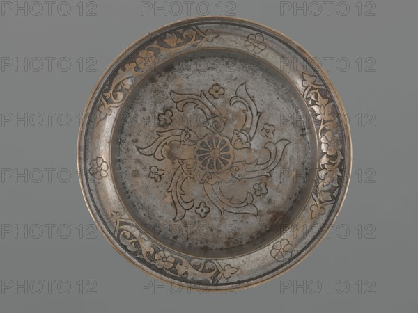 Bowl, Islamic art. Metal bowl with a flat bottom, low straight wall and slightly rounded rim. Decorated with palmettes on the outer walls, fish, floral ornament and rosettes. 18th century; possibly Turkey or Iran. Vessel; height 4.4 cm, diameter 21.2 cm., A round metal plate with a decorative floral pattern fills the frame, showing a central rosette surrounded by stylized petal and vine motifs; the rim has a band of engraved floral and scrolling designs. The surface shows areas of tarnish and wear with mottled textures. Visible colors include silver-gray of the metal, darker gray and black in the engraved lines and spots, and warm brownish-gold tones along the raised edges and decorative details.