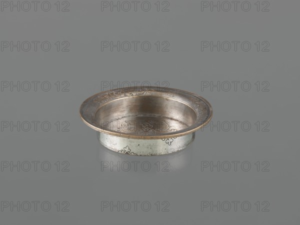 Bowl, Islamic art, metal bowl with fish, floral ornament and rosettes; flat bottom, low straight wall with slightly rounded rim; decorated with palmettes on the outer walls; 18th century; Turkey, / Iran ; vessel; height 4.4 cm, diameter 21.2 cm, A shallow round metal dish with a wide flared rim sits on a smooth gray surface, the dish showing a metallic palette of silver and warm bronze tones with subtle darker patina. The interior and rim display engraved floral and scroll motifs, and the exterior side bears small decorative patterns. The dish casts a soft reflection on the surface beneath it, and the overall scene contains shades of gray in the background and surface.