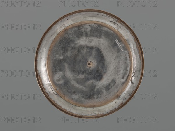 Bowl, Bowl with a flat bottom, low straight wall and slightly rounded rim; decorated with palmettes on the outer walls, fish, floral ornament, and rosettes. Metal. 18th century, possibly Turkey or Iran. Vessel; height 4.4 cm, diameter 21.2 cm., A round metal object viewed from above, centered against a flat gray background, with a worn reflective surface showing a mixture of silvery gray and darker gray tones and areas of brownish tarnish along the rim and inner ring; the surface has visible scratches and scuffs, a small central raised element, and a few irregular dark spots and blemishes around the edge.