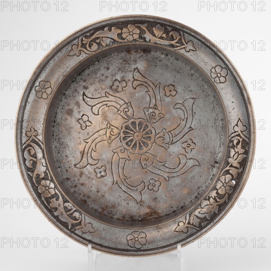 Bowl, Islamic art; metal bowl with flat bottom, low straight wall and slightly rounded rim; decorated with palmettes, fish, floral ornament, and rosettes on outer walls; 18th century; possibly Turkey or Iran; vessel; height 4.4 cm, diameter 21.2 cm., A round metal plate with a raised rim, showing a central floral medallion surrounded by swirling petal-like motifs and small flower shapes, all incised into the surface; the rim features a repeating decorative band of stylized flowers and vine patterns interspersed with plain floral motifs. The metal surface displays a mix of gray and brown tones with darker spots and patches of tarnish, and subtle highlights of a warm bronze or copper color along the raised designs and edges. The plate sits on a transparent stand against a neutral light background.