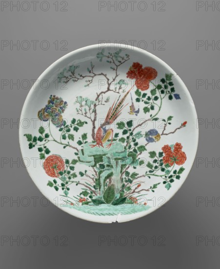 Platter, decorated with pheasants and flowers in colored porcelain, Qing dynasty 1644–1911, 18th–19th century, China; vessel; height 5.9 cm, diameter 35.4 cm., A round ceramic plate with a white background decorated with a painted scene in green, red, orange, yellow, blue, brown, and black; the central motif shows a stylized green rock formation with leafy green plants at its base and a colorful bird perched on top with long red and yellow tail feathers, surrounded by branches with green leaves and several large red-orange and multicolored blossoms, small buds, and scattered foliage, all rendered in fine brushwork against a neutral gray backdrop.