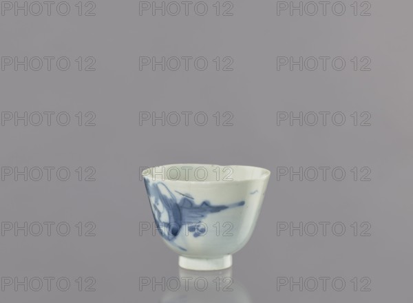 Bowl, Japanese, bowls, landscape motifs, ceramics, ca. 1750–1800, Japan place of origin?, vessel, height 6.3 cm, diameter 8.3 cm, A small ceramic cup with a pale off-white base and blue painted decoration sits centered against a smooth gray background. The cup has a slightly flared rim and a short, stepped foot; its surface shows a few irregularities and subtle gloss. The blue decoration appears as flowing brushstrokes with darker and lighter blue tones concentrated on one side, creating abstract shapes and small rounded marks. The interior of the cup is the same off-white color with faint blue tints near the rim. The scene includes reflections of the cup on the glossy surface beneath it. The visible colors are gray, off-white, and various shades of blue.