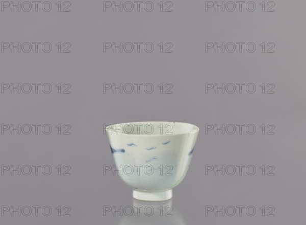 Bowl, Japanese, landscape motifs, ceramics, ca. 1750–1800, Japan, vessel; height 6.3 cm, diameter 8.3 cm, A small ceramic cup sits centered against a smooth gray background, the cup showing a pale off-white glaze with subtle blue brushstroke markings around its exterior, a few darker blue spots near the rim, a rounded body that tapers to a short foot, and a slight reflection visible on the surface beneath it; visible colors include gray, off-white, pale blue, and darker blue.