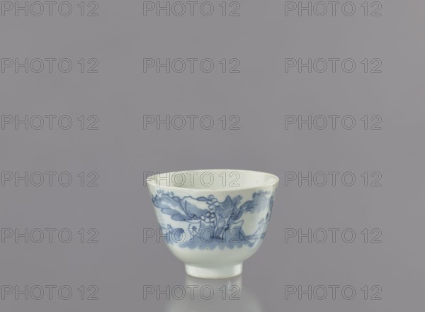 Bowl, Japanese porcelain bowl, widening toward the top, on a 0.5 cm foot. Decorated with a seaside landscape of fishermen among hills and coastal trees; two human figures—one with a fishing rod and the other with a net. Signature, on the bottom. Ca. 1750–1800, Arita, Japan. Vessel: height 6.3 cm, diameter 8.3 cm., A small porcelain cup sits centered against a uniform gray background, showing a pale off-white body with blue painted decorative motifs around its exterior; the cup has a rounded shape that tapers to a short foot, and the blue decoration includes flowing brushstrokes and floral or organic shapes in varying blue tones, with a smooth glossy surface that reflects light and casts a subtle reflection below.