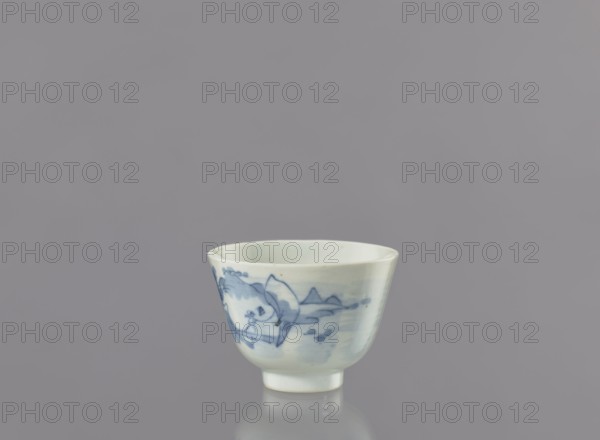 Bowl, Japanese art; porcelain bowl widening toward the top on a 0.5 cm foot. Decorated with a seaside landscape of fishermen among hills and coastal trees, showing two figures—one with a fishing rod and one with a net. Possible signature on the bottom. Ca. 1750–1800, Arita, Japan. Vessel height 6.3 cm, diameter 8.3 cm., A small ceramic cup sits on a reflective surface against a plain gray background, the cup predominantly white with blue painted designs suggesting natural forms; the interior is pale white, the rim slightly irregular, and the cup stands on a short foot with a soft shadow and reflection beneath it.