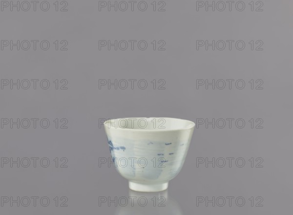 Bowl, Japanese art, Japanese culture; subject: fishermen, seaside landscape; object: porcelain bowl widening toward the top on a 0.5 cm foot; decorated with a view of fishermen on the seashore among hills and coastal trees, two human figures—one with a fishing rod and the other with a net; possible signature on the bottom; ca. 1750–1800, Arita, Japan; vessel height 6.3 cm, diameter 8.3 cm; unknown provenance., A small porcelain cup sits centered against a plain gray background, showing a smooth off-white surface with subtle pale blue brushlike marks and a few darker blue spots; the cup has a slightly flared rim, a rounded body on a short foot, and a soft reflective shadow beneath it.