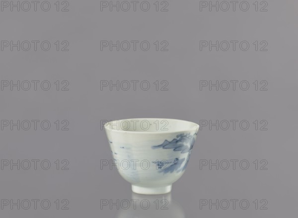 Bowl, Japanese, fishermen, seaside landscapes, bowls, landscape motifs, ceramics, unknown provenance. Porcelain bowl widening towards the top, on a 0.5 cm foot. Decorated with a view of fishermen on the seashore among hills and coastal trees; two human figures: one with a fishing rod and the other with a net. Signature, on the bottom. Ca. 1750–1800, Arita, Japan. Vessel height 6.3 cm, diameter 8.3 cm., A small ceramic cup sits centered against a smooth gray background, showing a pale off-white body with delicate blue-painted motifs on its exterior; the cup has a slightly flared rim, a rounded bowl shape narrowing to a short foot, and faint reflections on the surface beneath it.